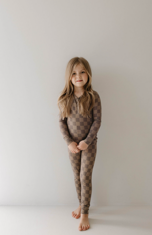 A young girl with long hair stands against a white wall. She is barefoot and dressed in hypoallergenic baby clothing from forever french baby, wearing the Bamboo Two Piece Pajama in Faded Brown Checkerboard. She smiles softly, with her hands clasped in front of her.
