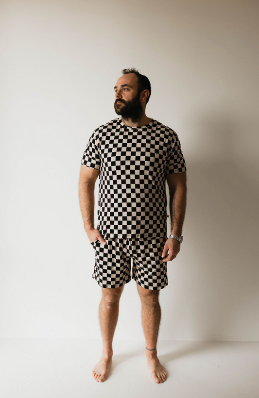 A man stands barefoot against a plain white background, wearing the forever french baby Adult Bamboo Short Pajamas in Black Checkerboard. He is looking off to the side, with his hands relaxed by his sides. He has a beard and a watch on his left wrist.