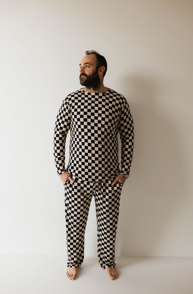 A bearded man with a mustache stands barefoot against a white background, donning the Adult Bamboo Pajamas in a black-and-white checkerboard pattern from Forever French Baby. He looks off to the side with his hands in his pockets, exuding style reminiscent of the brand's offerings.
