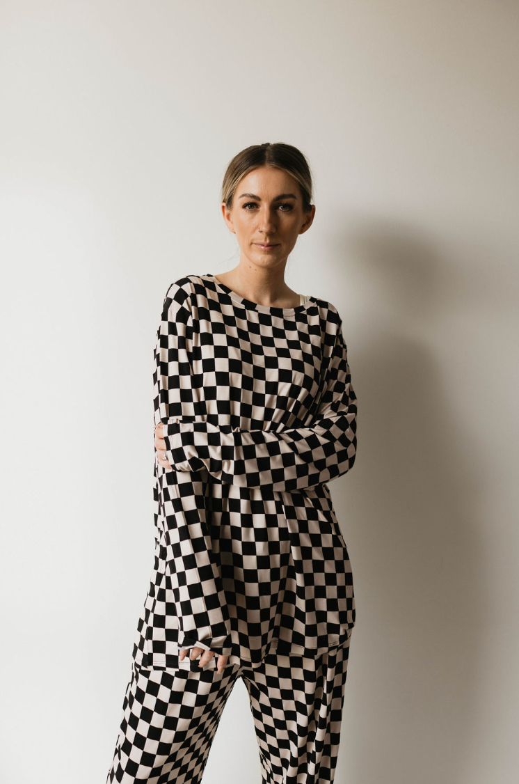 A woman stands against a plain white background, wearing the Adult Bamboo Pajamas in a black and white checkerboard pattern by forever french baby. She has her hair pulled back and is staring directly at the camera with a neutral expression, one arm crossed over her body.