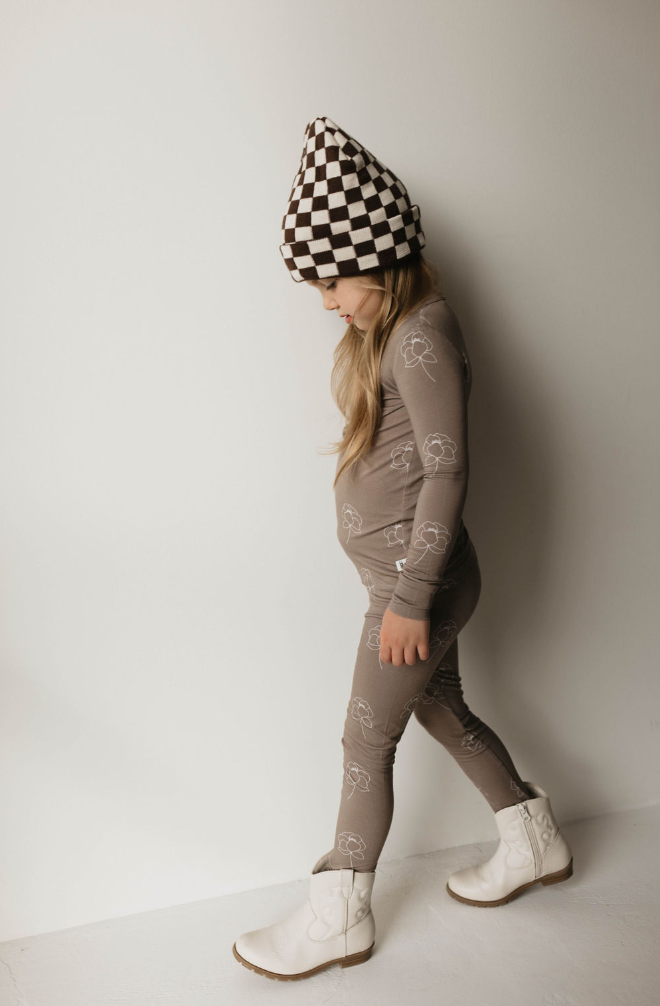 A young girl wearing forever french baby's premium Bamboo Two Piece Pajama | Sweet Dreams Floral in a brown outfit with white patterns, white boots, and a brown and white checkered hat walks against a plain white background. She is looking down and has long hair flowing from under her hat. Her hypo-allergenic clothing ensures comfort and style.