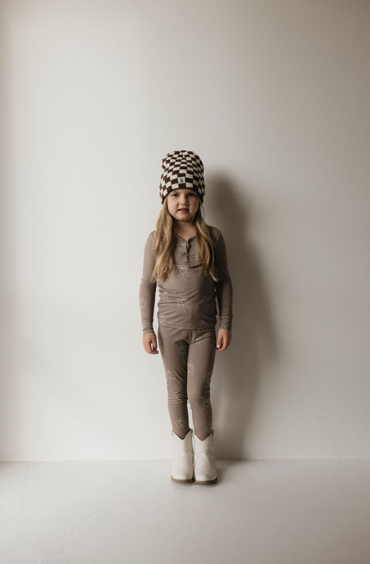 A young child with long hair stands against a plain white wall. They are wearing a brown, long-sleeve top and matching pants made from the "Bamboo Two Piece Pajama | Sweet Dreams Floral" by forever french baby, complemented by a black and white checkered beanie, and white boots. The child's expression is neutral, looking directly at the camera.