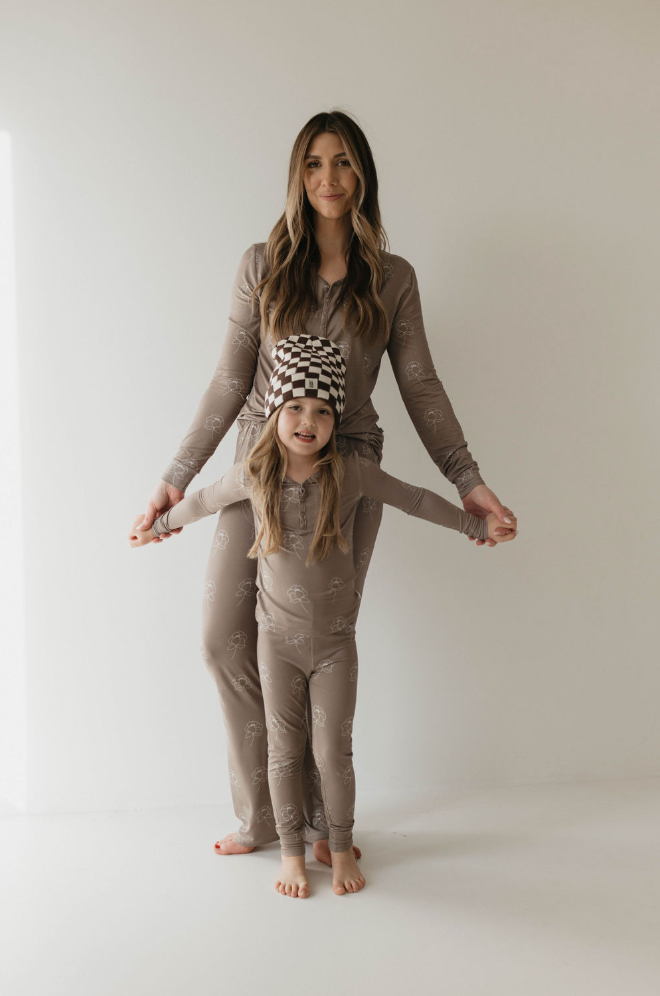 A woman and a child, both with long hair, stand against a plain white backdrop wearing matching Sweet Dreams Floral bamboo pajamas from Forever French Baby. The child wears a brown and white checkered beanie, standing with arms outstretched while holding the woman's hands.