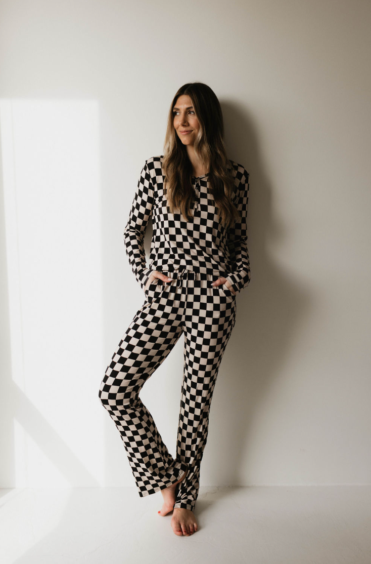 A woman with long hair stands barefoot against a white wall, wearing Women's Bamboo Pajamas in Black Checkerboard by forever french baby. With her hands in her pockets, she looks slightly to the side and smiles softly as sunlight casts a shadow on the wall next to her.