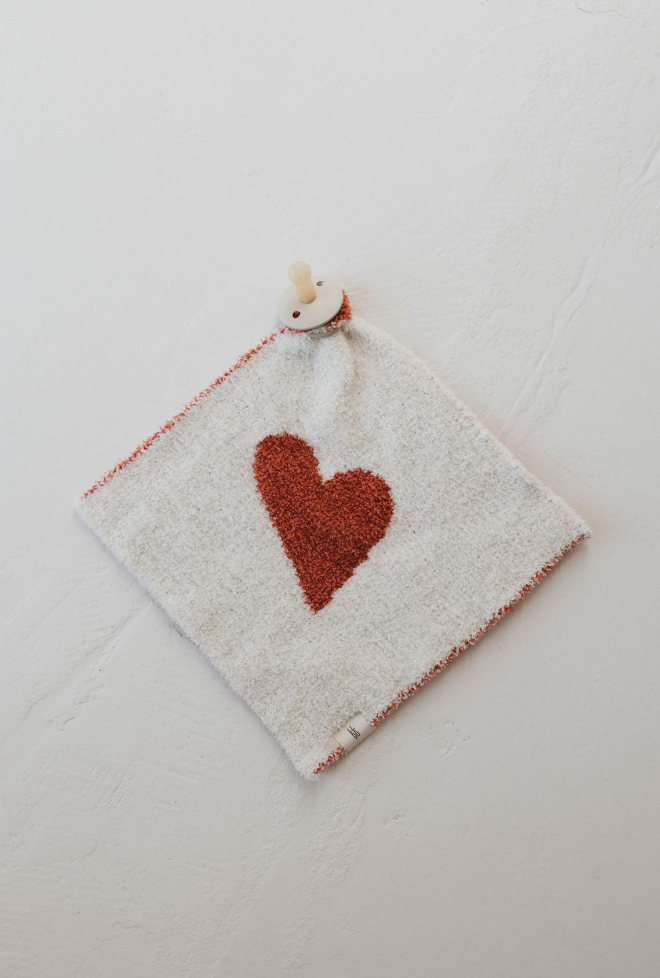 The "Lovey | Queen of Hearts" by forever french baby features a soft, white microfiber feather yarn cloth that is neatly folded and decorated with a rust-colored heart in the center. A small wooden clip adorns one corner of this elegant lovey. The textured surface and refined edging lie flat against a light, textured background.