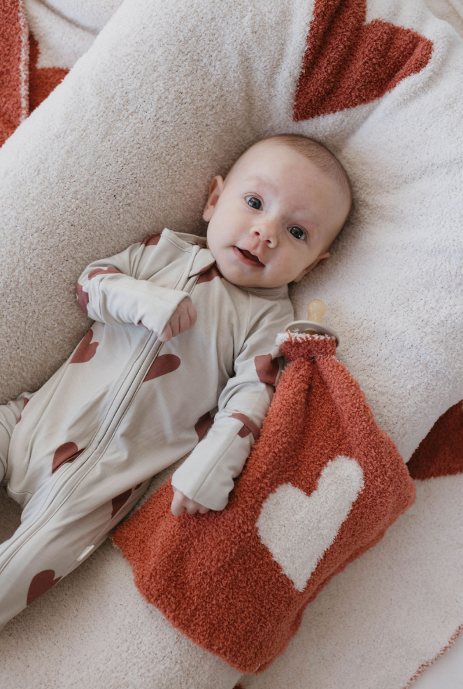 A baby lies on a white and red blanket with heart patterns. The baby, adorned in a light-colored onesie with red heart designs, clutches the "Lovey | Queen of Hearts" from the forever french baby collection. Gazing upward with a calm expression, the scene exudes pure sweetness.