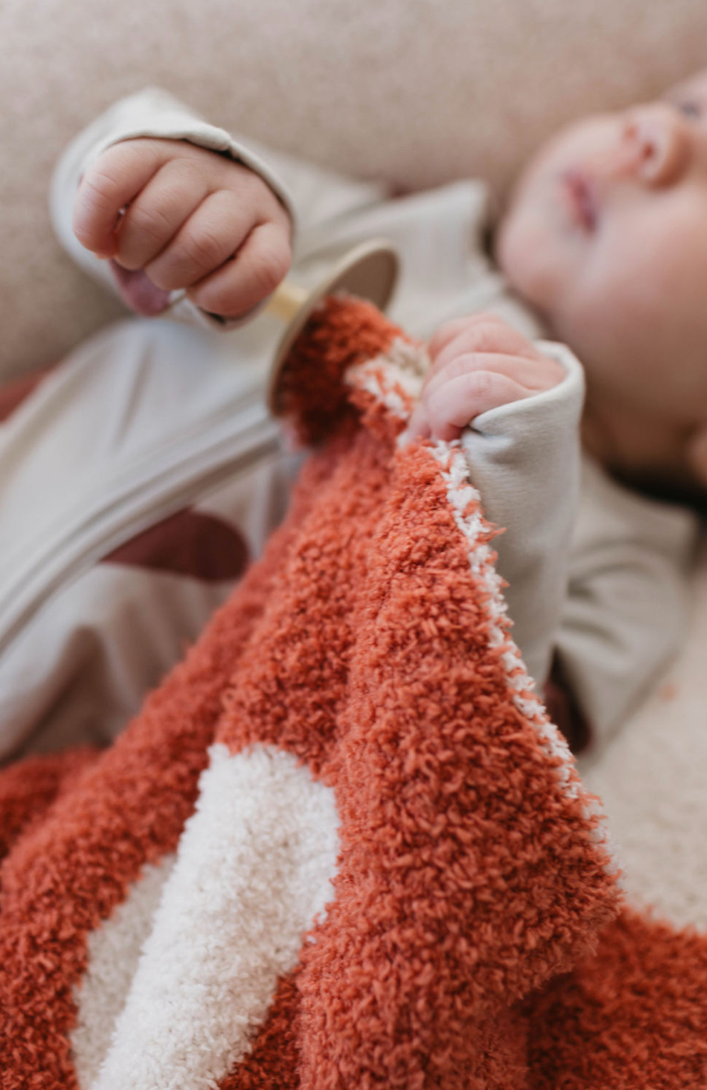 A baby in a grey outfit tightly holds the Queen of Hearts lovey by forever french baby, a soft and fuzzy red and white blanket made of microfiber feather yarn, while lying down. The baby's hands clutch the edge of the lovey, showcasing the texture and warmth of the fabric.