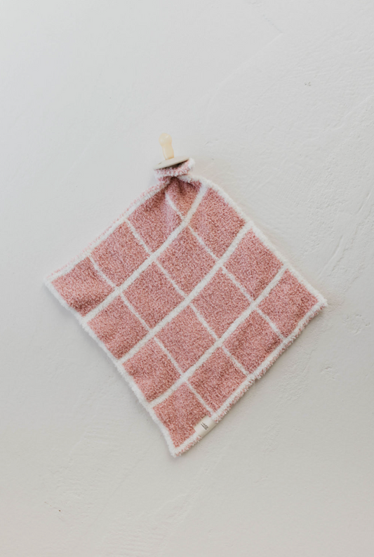 A Lovey | Cotton Candy Grid cloth hangs on a peg against a white textured wall. Crafted from the softest lovey microfiber feather yarn by forever french baby, the pink and white checkered cloth has a fluffy texture and is bordered in pristine white.