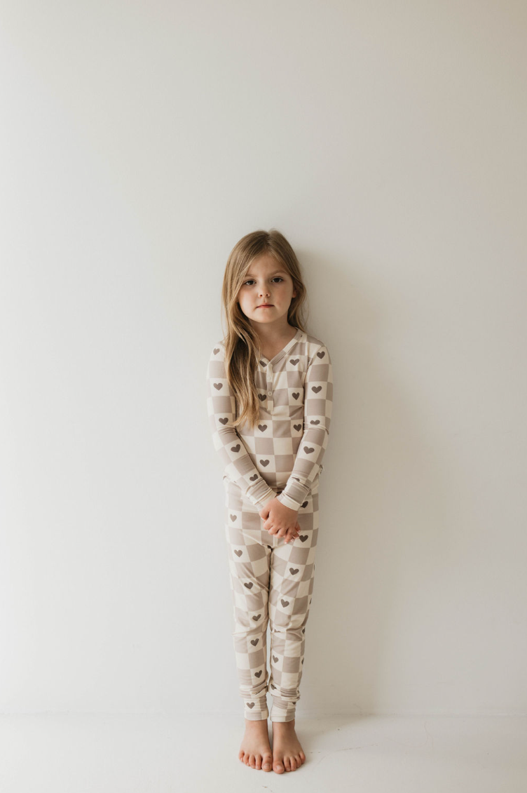 A young child with long, brown hair is standing against a plain white wall. They are wearing the forever french baby Bamboo Two Piece Pajama from the Check Your Heart collection, featuring a beige and white checkered pattern adorned with brown heart shapes. The child is barefoot and has a slight, serious expression on their face, arms relaxed at their sides.