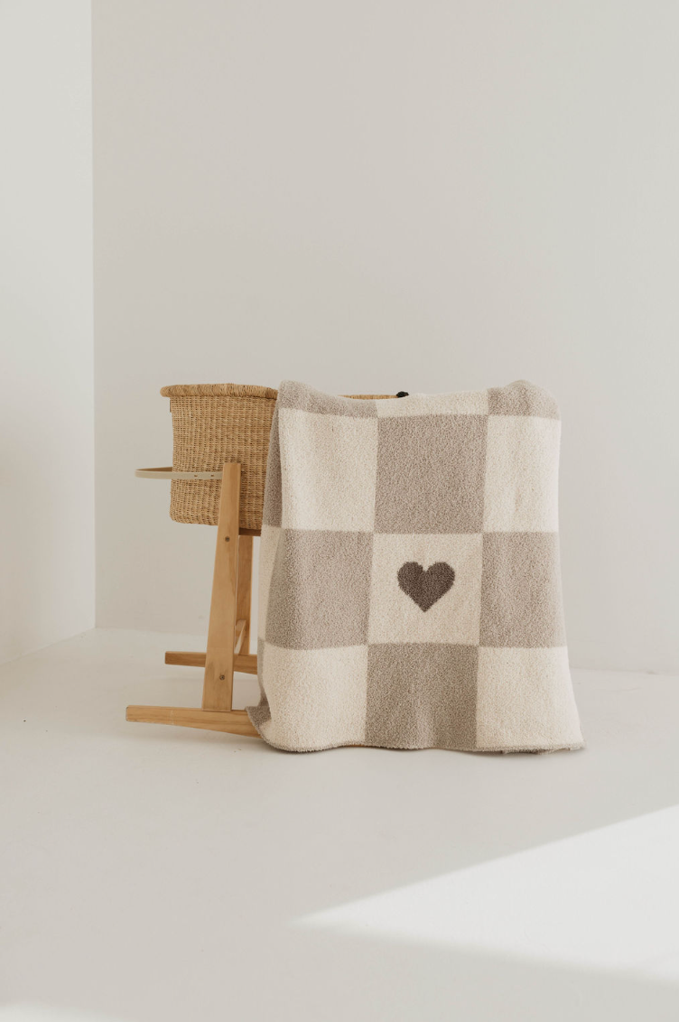 A wooden stand holds a woven basket, partially draped with the Plush Blanket | Check Your Heart by forever french baby, featuring cozy checkered neutral tones. This soft blanket, crafted from OEKO Standard 100 materials, showcases a heart pattern in its center. The minimalist setting is brightly lit, accentuated by light-colored walls and a clean, white floor.