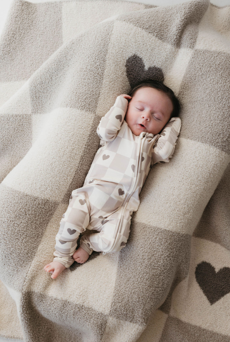 A baby sleeps peacefully on a cozy, "Check Your Heart" blanket by forever french baby, featuring heart patterns. Made from OEKO Standard 100 materials, the plush blanket complements the matching onesie adorned with hearts. The baby's hands are positioned near their head, and the colors are in soft, neutral tones of beige and gray.
