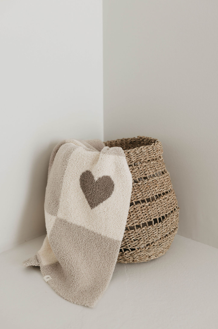 A woven basket is placed in a corner, with a Plush Blanket | Check Your Heart by forever french baby draped over its side. The blanket, made from OEKO Standard 100 materials, features a dark heart pattern on one of its squares. The scene is minimalistic and serene, with neutral colors and simple textures.