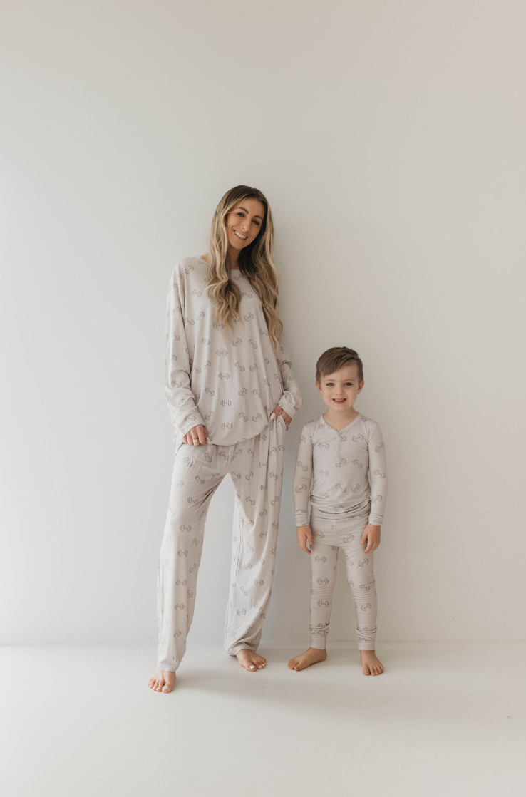 A woman and a young boy stand against a plain white background, both wearing matching light-colored Adult Long Sleeve Bamboo Pajamas by forever french baby with a subtle pattern. The woman, with long hair, smiles gently, while the boy stands close to her, smiling with one hand in his pocket, ready for a comfortable night's sleep.