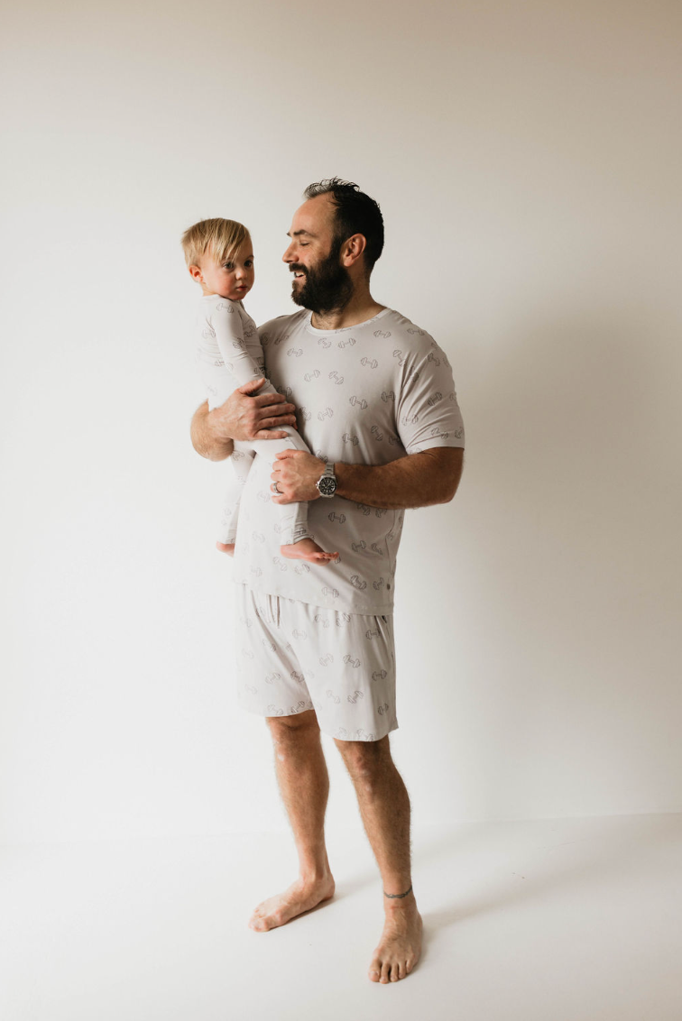 In a white room, a man stands barefoot holding a young child. Both are dressed in matching light-colored "Adult Bamboo Short Pajamas | Arm Day" from forever french baby, featuring a subtle dumbbell weights graphic. The man gazes at the child with a smile, exuding warmth and familial affection.