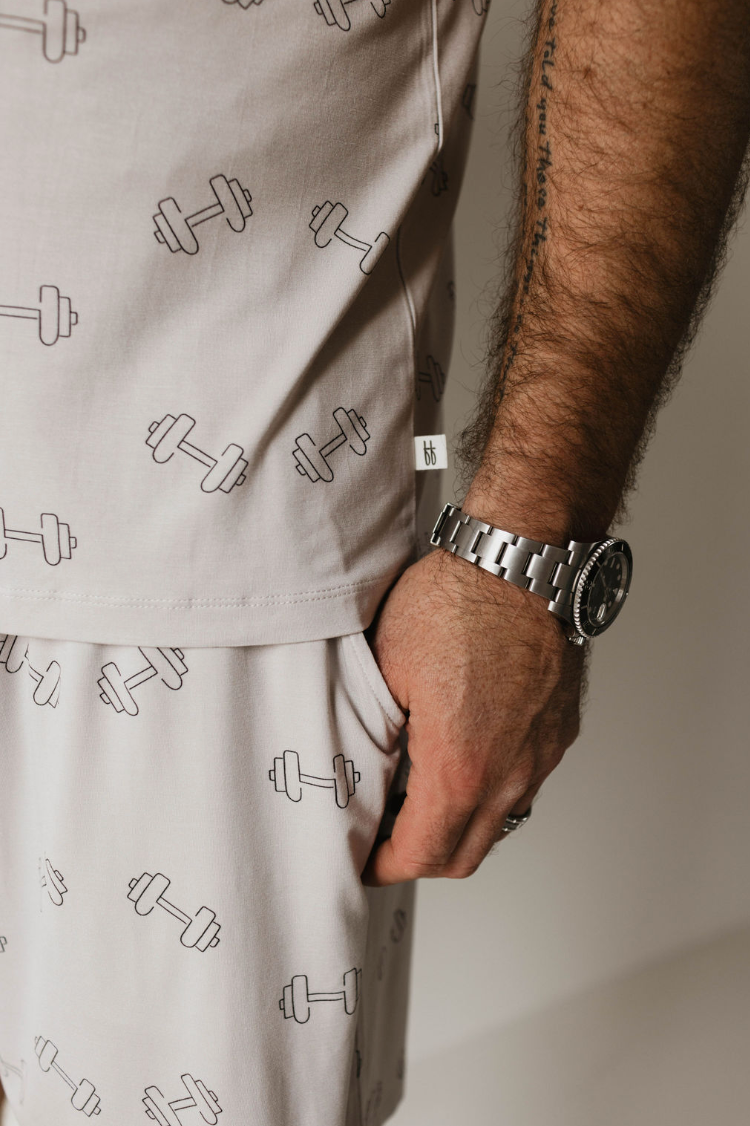 Close-up of a person wearing light-colored Adult Bamboo Short Pajamas in the "Arm Day" design by forever french baby, featuring a dumbbell weights graphic. The person’s right hand is partially inside a pocket, showcasing a wristwatch and a tattoo with delicate handwriting on their forearm. The background is neutral and out of focus.