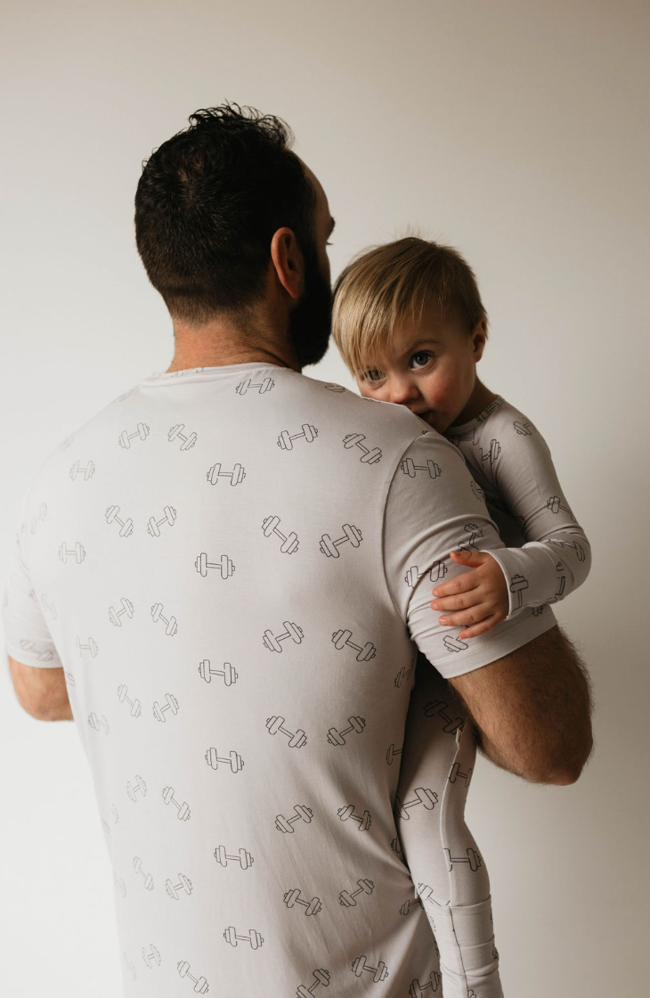 A man with short dark hair, viewed from behind, holds a young child. Both are sporting matching light grey forever french baby Adult Bamboo Short Pajamas featuring a fun dumbbell weights graphic from the Arm Day collection. The blond-haired child is facing the camera while resting their head on the man's shoulder.