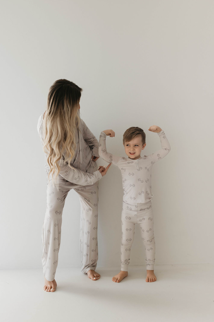 An adult and a child wearing matching Adult Long Sleeve Bamboo Pajamas | Arm Day by forever french baby stand against a plain white background. The child faces the camera, smiling, and flexing their arms proudly while the adult, standing next to them, looks towards the child. Both sets of pajamas feature a subtle dumbbell weights graphic pattern.