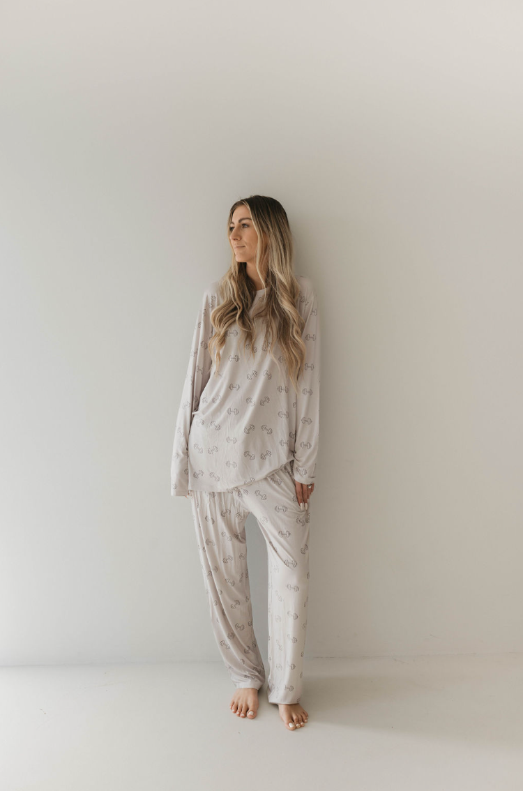 A woman with long hair stands barefoot against a blank wall, wearing light-colored **Adult Long Sleeve Bamboo Pajamas | Arm Day** by *forever french baby* featuring a simple pattern. Her hands are by her sides, and she gazes off to the left with a calm expression, suggesting she's ready for a comfortable night's sleep.