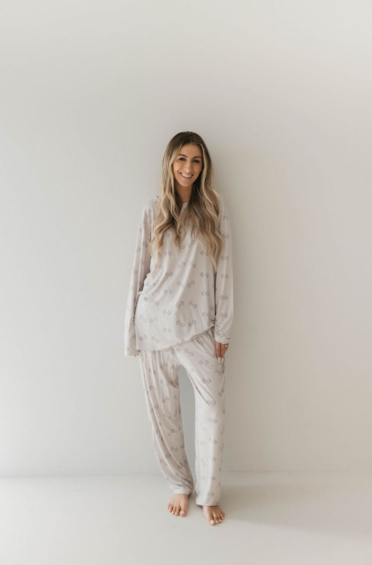 A woman with long, wavy hair stands against a white background, smiling. She is wearing a loose, light-colored pajama set from forever french baby’s Adult Long Sleeve Bamboo Pajamas in the "Arm Day" style, featuring a subtle pattern. She is barefoot and appears relaxed and comfortable, ready for a restful night's sleep.