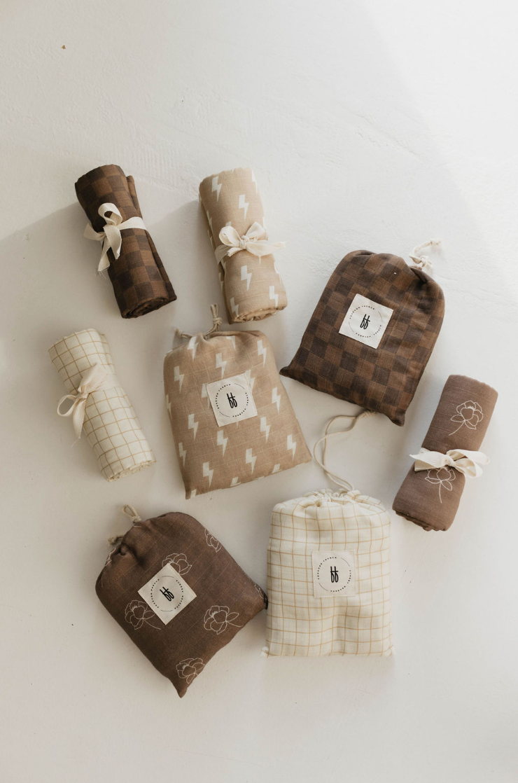 Nine rolled and bundled fabric swatches are arranged on a white surface. The fabrics, showcasing patterns such as checkered, floral, grid, and lightning bolts, come in shades of brown, beige, and cream. Some bundles are tied with cream-colored strings. Among them is the **Golden Grid Muslin Swaddle by forever french baby**.