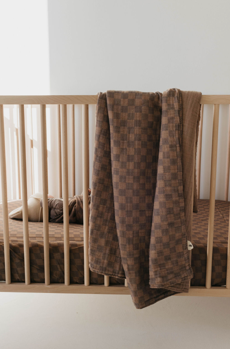 A minimalist wooden crib with wooden slats is adorned with the Muslin Crib Sheet | Faded Brown Checkerboard from forever french baby. Inside, there is a nursery bedding set featuring this soft muslin cotton crib sheet and a stuffed toy. Draped over the side of the crib is a matching checkered brown blanket. Soft natural light filters through the room.