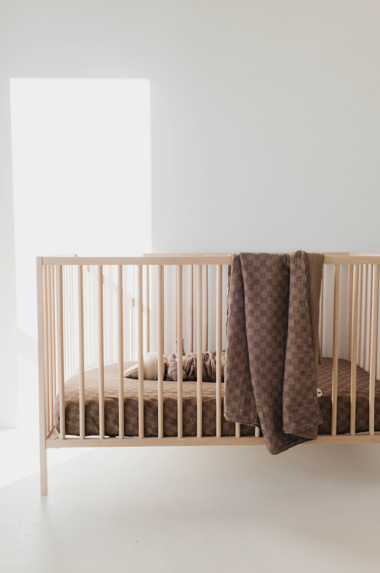 A simple wooden baby crib stands in a sunlit room with a white wall. A Muslin Crib Sheet in Faded Brown Checkerboard by forever french baby is draped over the side, and nursery bedding with a small stuffed animal are placed inside the crib. The area is minimal and serene.
