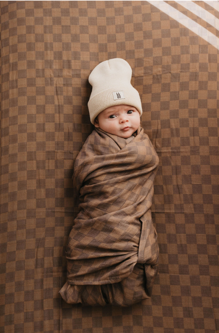 A baby, wrapped snugly in a brown checkered blanket, lies on a matching Muslin Crib Sheet | Faded Brown Checkerboard by forever french baby. Wearing a light beige knit beanie and looking slightly off-camera with a calm expression, the serene scene showcases the softness of muslin cotton nursery bedding.