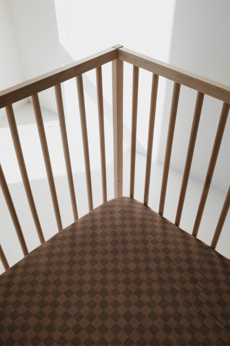 An empty wooden crib with vertical slats on a sunlit floor features the "Muslin Crib Sheet | Faded Brown Checkerboard" by forever french baby. Soft sunlight casts shadows through the slats, creating a calm and peaceful atmosphere perfect for nursery bedding in muslin cotton.