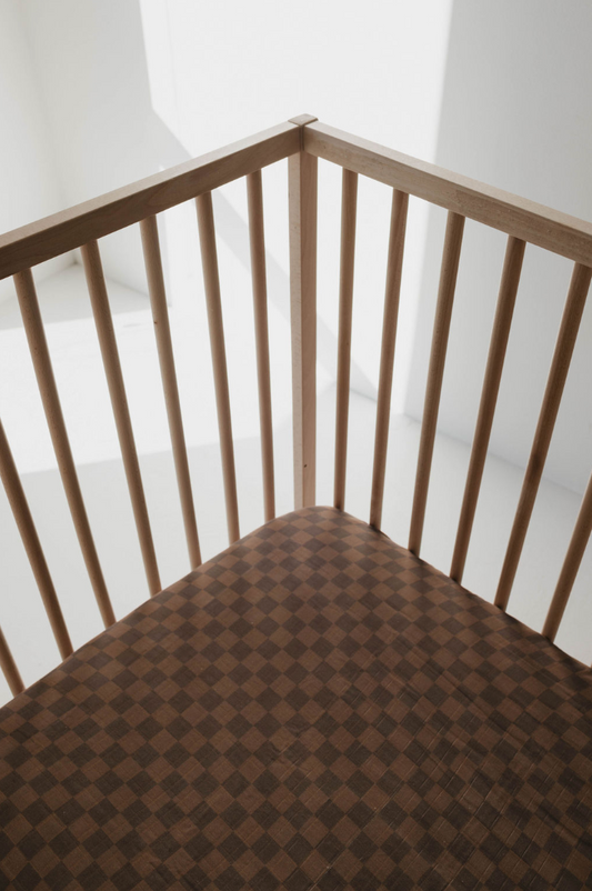 An empty wooden crib with vertical slats on a sunlit floor features the "Muslin Crib Sheet | Faded Brown Checkerboard" by forever french baby. Soft sunlight casts shadows through the slats, creating a calm and peaceful atmosphere perfect for nursery bedding in muslin cotton.