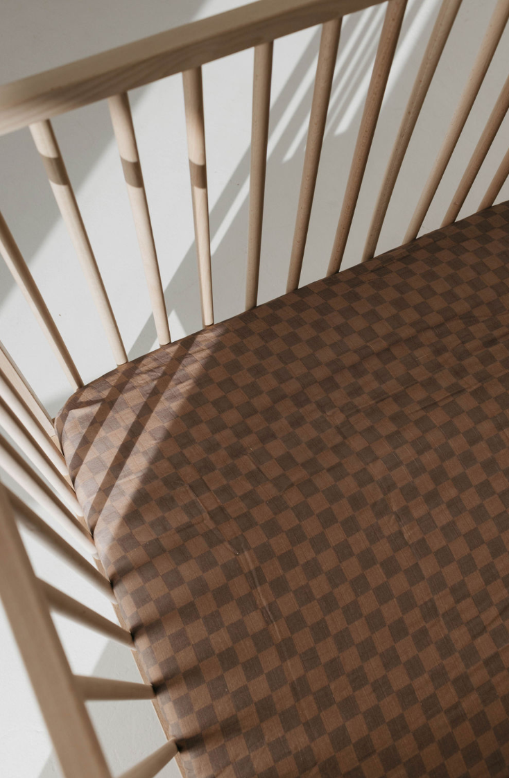 A top-view of a wooden crib with vertical slats shows the mattress covered with the Muslin Crib Sheet | Faded Brown Checkerboard by forever french baby, made from soft muslin cotton. Sunlight casts soft shadows through the slats onto the mattress, while the light-colored floor enhances the serene nursery bedding.