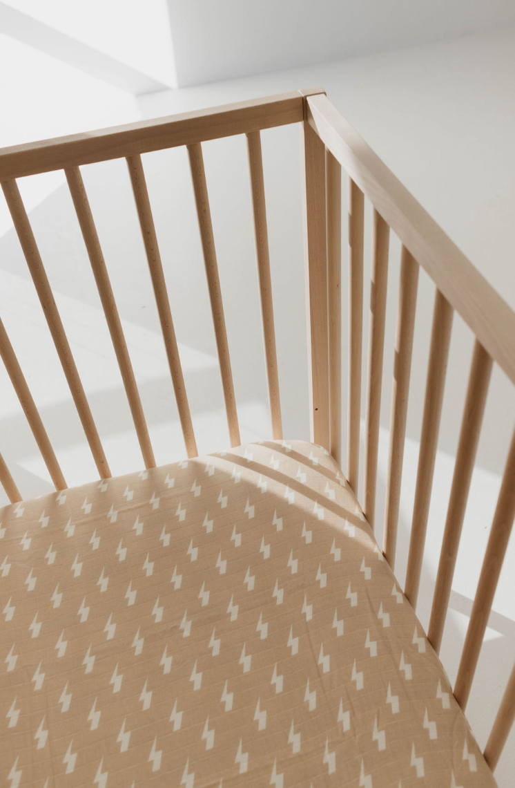 A corner of a wooden baby crib with vertical slats is shown. The crib mattress is adorned with the forever french baby Muslin Crib Sheet | Tan & Cream Lightning Bolt, crafted from soft muslin cotton. Gentle natural light enters from the left side, casting delicate shadows on this piece of modern nursery bedding.