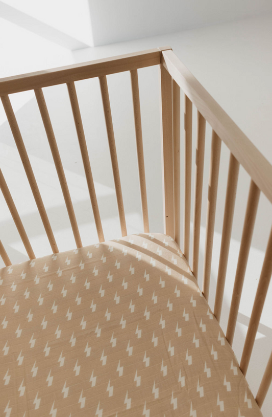 A corner of a wooden baby crib with vertical slats is shown. The crib mattress is adorned with the forever french baby Muslin Crib Sheet | Tan & Cream Lightning Bolt, crafted from soft muslin cotton. Gentle natural light enters from the left side, casting delicate shadows on this piece of modern nursery bedding.