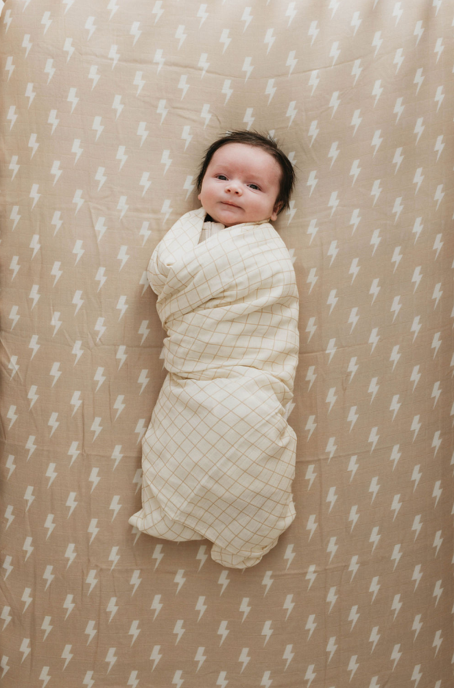 A baby lies in a crib, swaddled in a cream-colored blanket with a grid pattern. The muslin cotton crib sheet beneath the baby is adorned with white lightning bolts on a beige background. The Muslin Crib Sheet | Tan & Cream Lightning Bolt from forever french baby complements the baby's slight smile and dark hair perfectly.