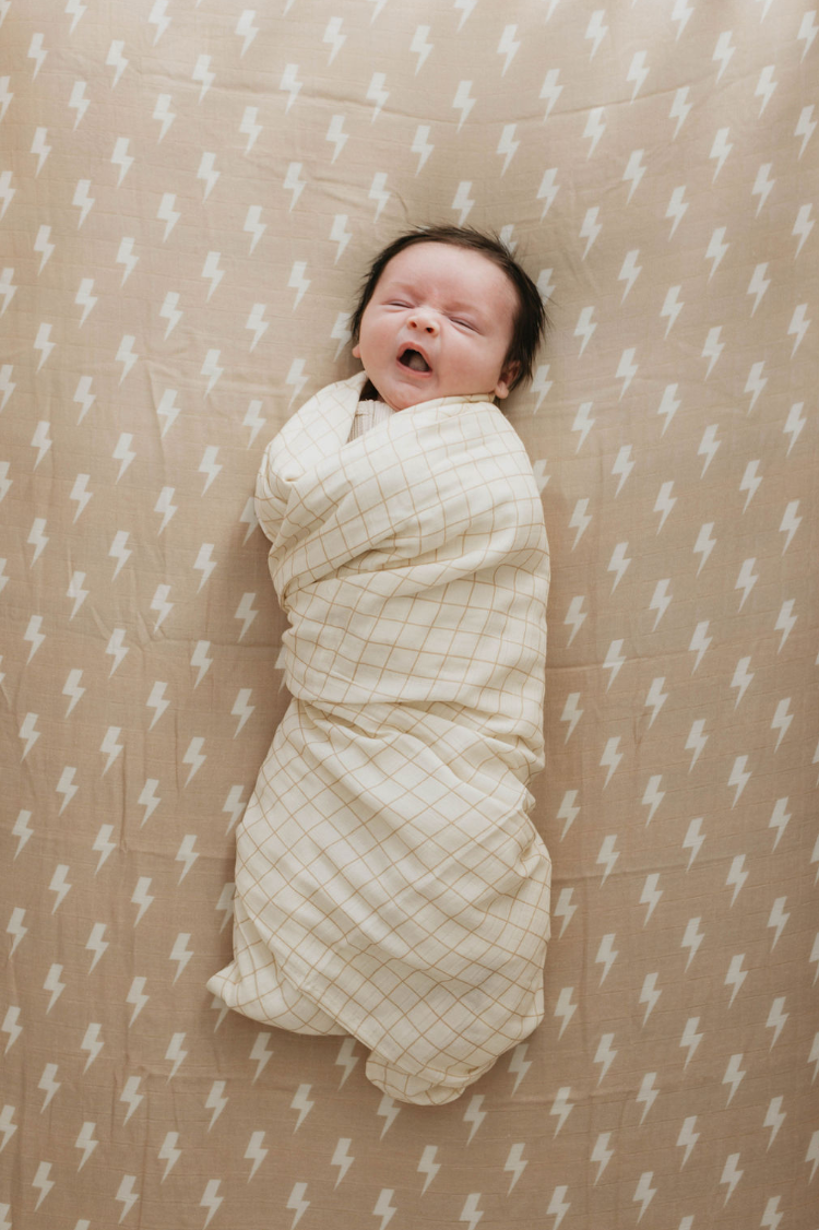 A baby swaddled in a beige and white checkered blanket lies on a *Muslin Crib Sheet | Tan & Cream Lightning Bolt* by forever french baby. The pale brown muslin cotton crib sheet features a striking white lightning bolt pattern. The baby, with dark hair, appears to be yawning or stretching, creating a serene moment in this modern nursery bedding setup.