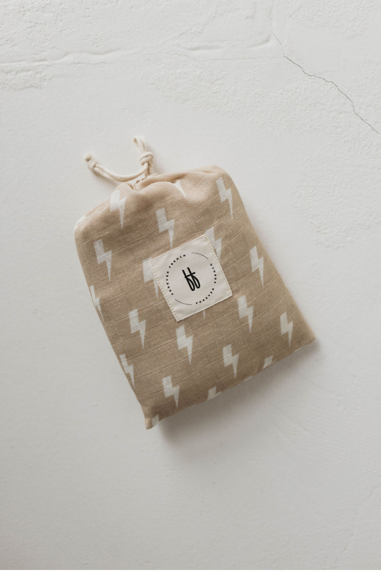 A petite beige drawstring bag adorned with a white lightning bolt pattern, echoing the design of modern nursery bedding like the Muslin Crib Sheet in Tan & Cream Lightning Bolt from forever french baby, and featuring a circular white tag with a minimalist black logo at its center. The bag is placed on a textured light-colored surface.