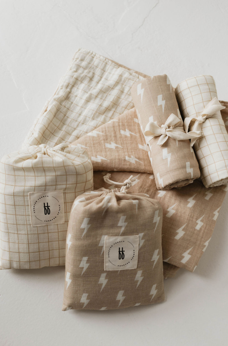 A flat lay of beige and white baby items showcasing various patterns, including lightning bolts and checkered designs. The collection features pouches, a blanket, and rolled-up cloths—all adorned with the "bb" logo on some pouches. The highlight is the Muslin Crib Sheet in Tan & Cream Lightning Bolt from forever french baby, adding a touch of softness to the modern nursery bedding collection.