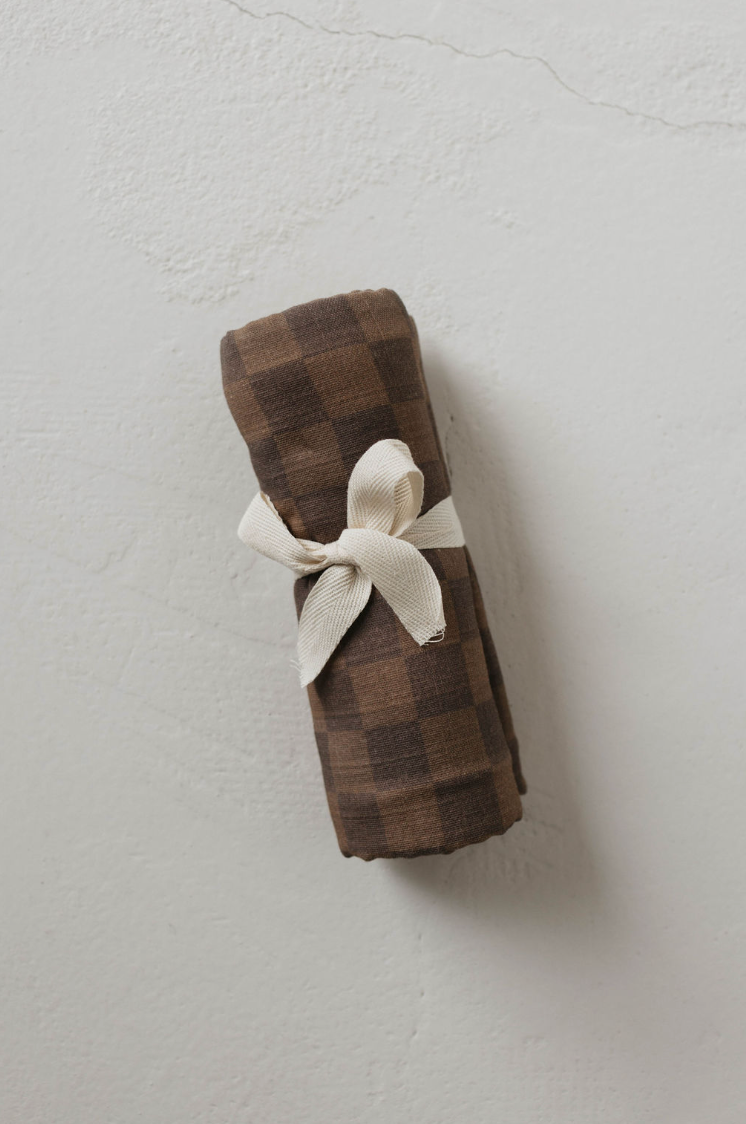 A neatly tied cream-colored ribbon holds the rolled-up forever french baby Muslin Swaddle | Faded Brown Checkerboard, set against a textured light gray background. The swaddle looks orderly and impeccably wrapped.
