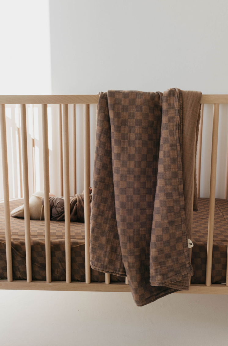 A wooden baby crib with a forever french baby Muslin Swaddle in Faded Brown Checkerboard draped over the side. Inside the crib, matching checkered bedding complements the swaddle, while a few soft toys rest on the mattress. Sunlight streams softly into the room, creating a play of light and shadows.