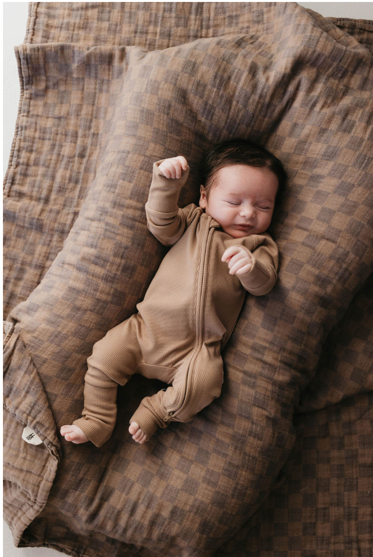 A baby, dressed in a brown onesie, sleeps peacefully on the forever french baby Muslin Quilt in the Faded Brown Checkerboard pattern. The soft muslin material of the quilt envelops the baby, giving the nursery a cozy and snug appearance as the baby raises both arms.