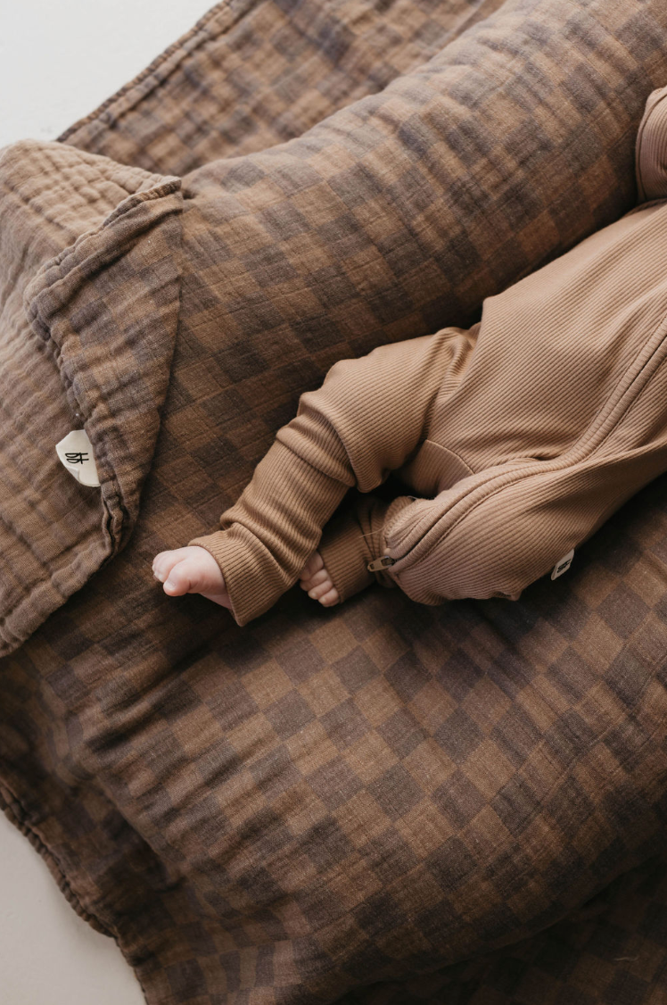 A baby swaddled in a brown blanket lies on a Muslin Quilt | Faded Brown Checkerboard by forever french baby. The baby's arm and foot, wearing a brown ribbed onesie, are visible. The quilt features button decorations and textured fabric, making it perfect for any baby nursery.
