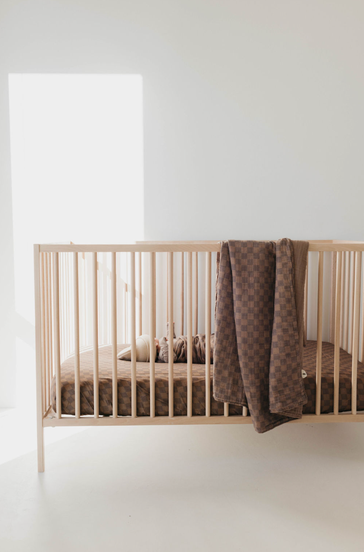 A minimalist light wooden baby crib is shown with a Muslin Quilt | Faded Brown Checkerboard by forever french baby draped over the railing. The crib's muslin bedding is brown, and it is positioned near a bright window in the baby nursery, with sunlight streaming in, creating a serene and cozy atmosphere.