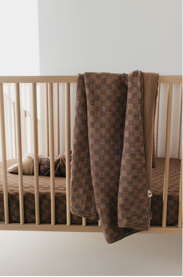 A cozy, minimalist baby nursery features a wooden crib with vertical slats and a Muslin Quilt in Faded Brown Checkerboard from forever french baby draped over the side. Inside the crib, a baby is lying on a matching muslin-sheeted mattress, partially covered by a soft brown blanket.
