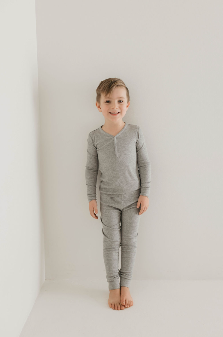 A young child stands barefoot against a white wall, smiling at the camera. Dressed in forever french baby's Bamboo Two Piece Pajamas in ribbed grey, the child radiates comfort and joy. This eco-friendly kids sleepwear enhances the minimalistic and bright atmosphere, emphasizing sustainability and style.