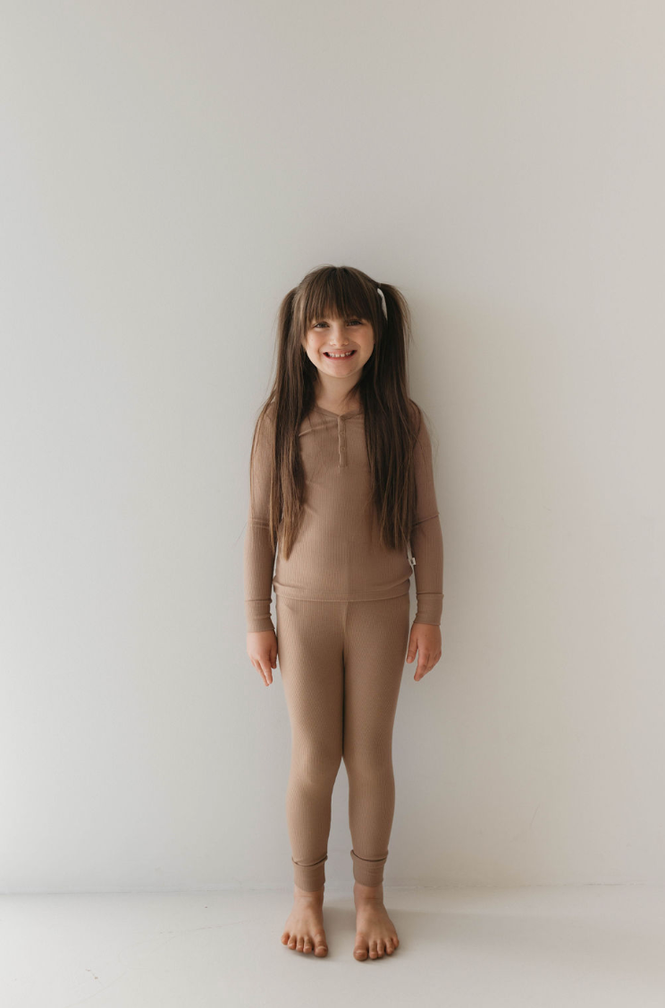 A young girl with long dark hair in pigtails stands barefoot against a white wall. She is wearing a matching "Two Piece Bamboo Pajamas" set in "Ribbed Milk Chocolate" by forever french baby. She appears to be smiling, showcasing the comfort of eco-friendly sleepwear for kids.