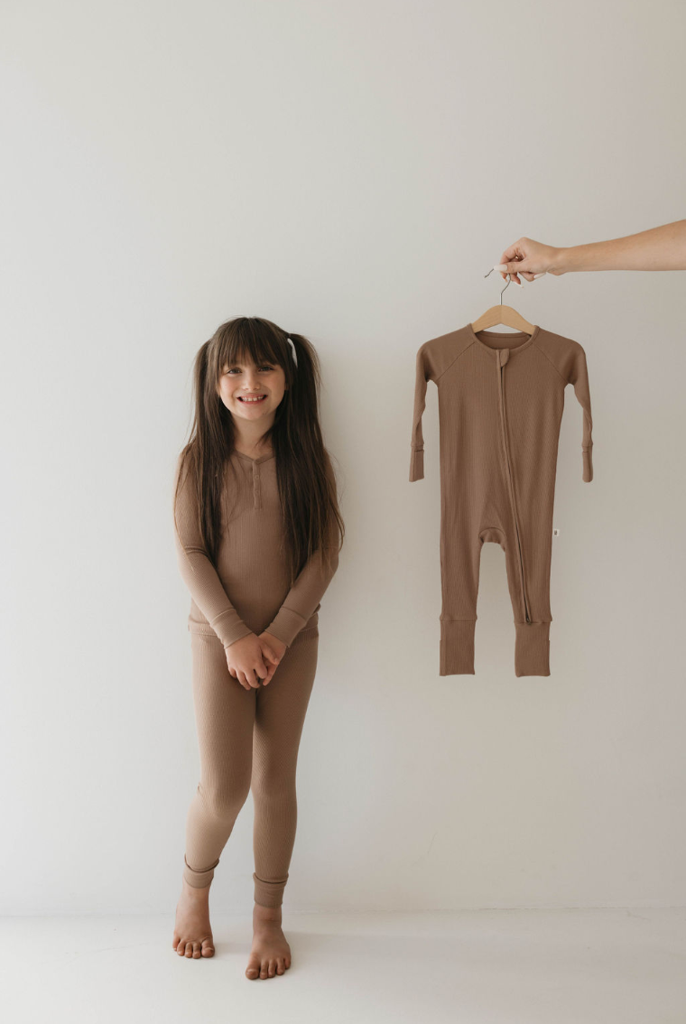 A young girl with long dark hair in pigtails, wearing a brown long-sleeve outfit, stands barefoot against a plain white background, smiling. Beside her, a hand holds up the "Two Piece Bamboo Pajamas | Ribbed Milk Chocolate" by forever french baby on a hanger—showcasing the comfort of eco-friendly sleepwear for kids.