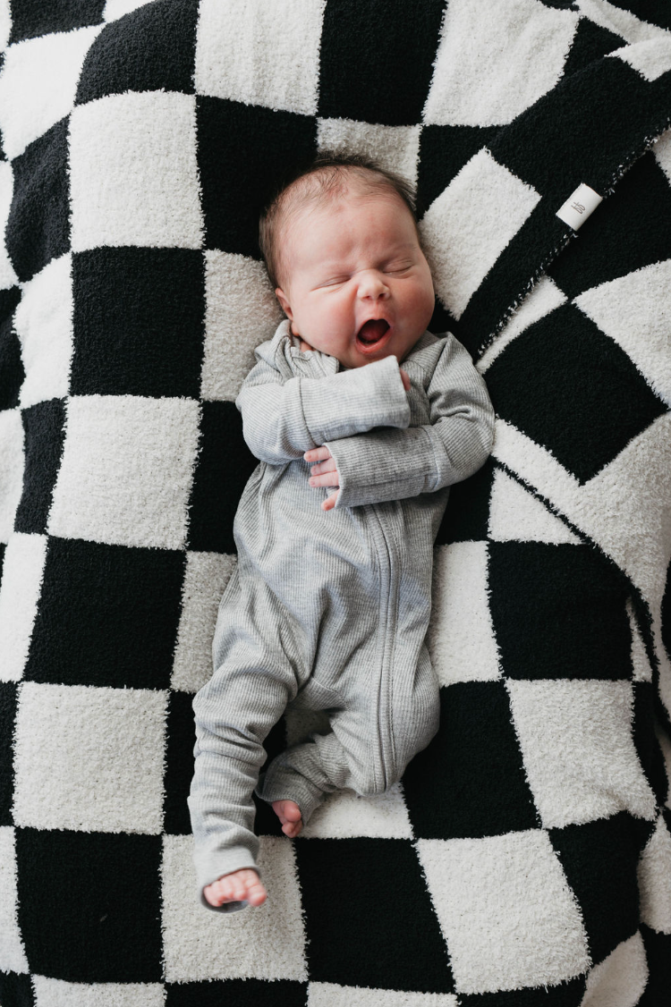 A baby wearing forever french baby's Ribbed Grey Bamboo Zip Pajamas yawns while lying on a black and white checkered blanket.