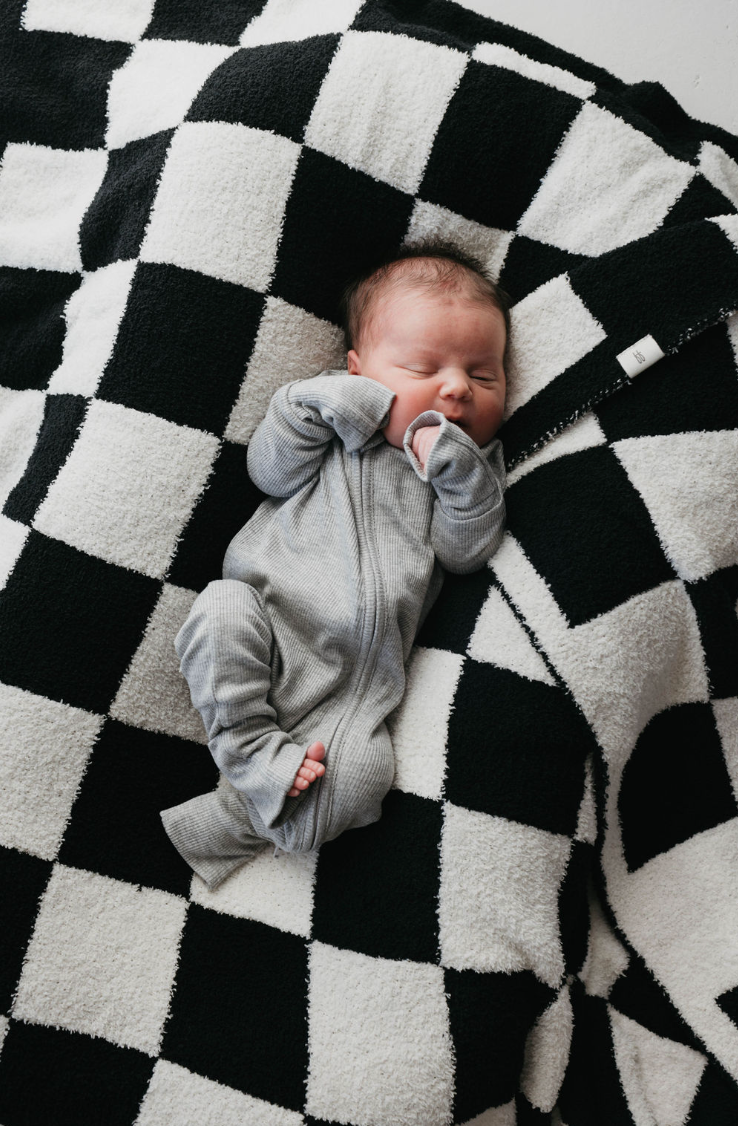 A baby in Bamboo Zip Pajamas (Ribbed Grey) by forever french baby rests peacefully on a black and white checkered blanket, surrounded by timeless style.