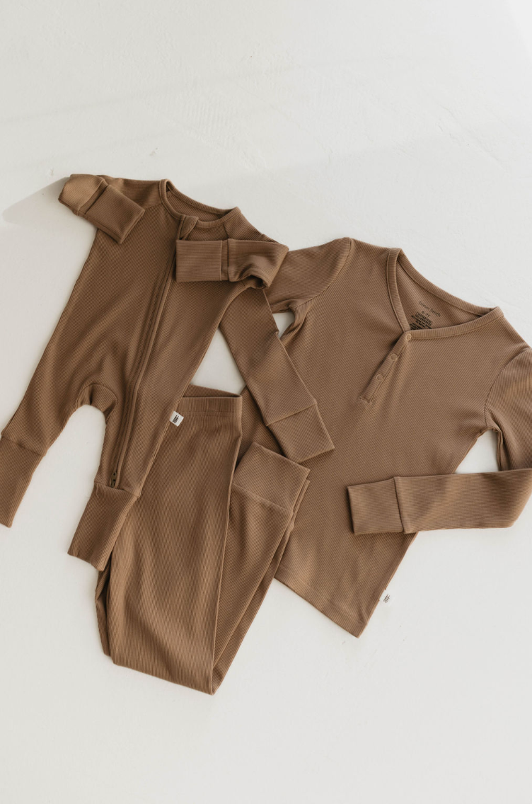 Three pieces of matching brown kids' loungewear from forever french baby are displayed on a white surface: a baby onesie with a zipper, a long-sleeve henley shirt, and a pair of pants. The eco-friendly sleepwear features ribbed bamboo fabric in the consistent "Milk Chocolate" color across all items.