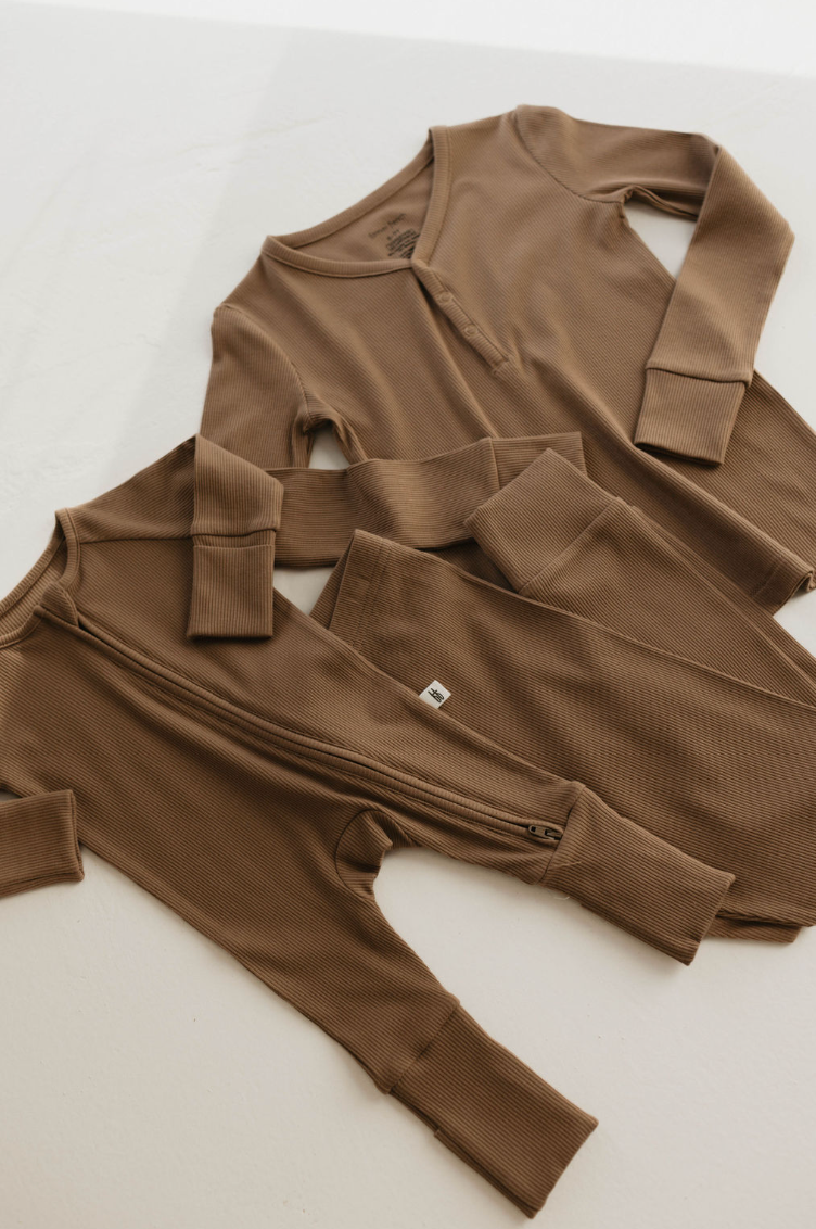 Two pieces of forever french baby's Ribbed Milk Chocolate bamboo pajamas are arranged on a white surface. One set features a button-up neckline, while the other has a full front zipper. Both sets include long sleeves and pants with ribbed cuffs. The fabric looks soft and cozy, making it ideal for eco-friendly kids' sleepwear.