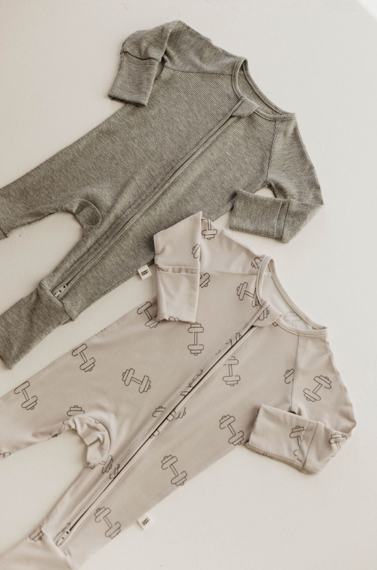 Two baby onesies are displayed. The top onesie, from Forever French Baby, is ribbed grey like the Bamboo Zip Pajamas; the bottom is light gray with a small dumbbell pattern. Both have front zippers and long sleeves.